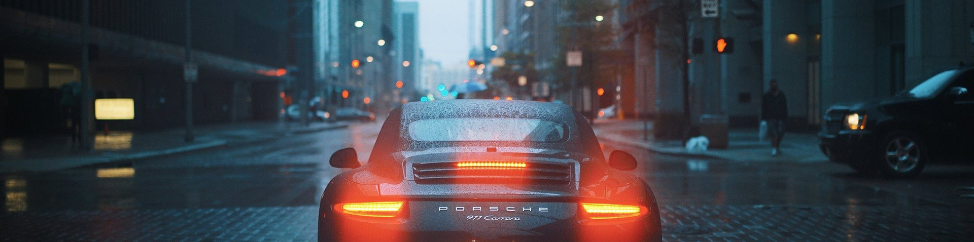 Porsche 911 Carrera von hinten mit leuchtenden Rücklichtern auf regennasser Stadtstraße – Porsche Success Story