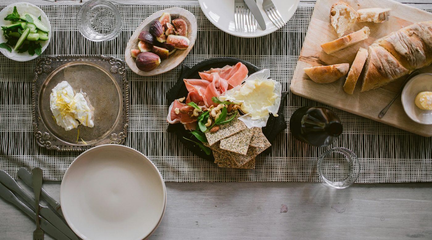 Gedeckter Tisch von oben mit Feigen, Rohschinken, Weichkäse, Knäckebrot und aufgeschnittenem Baguette – Produktdaten von Egle zentral in OMN