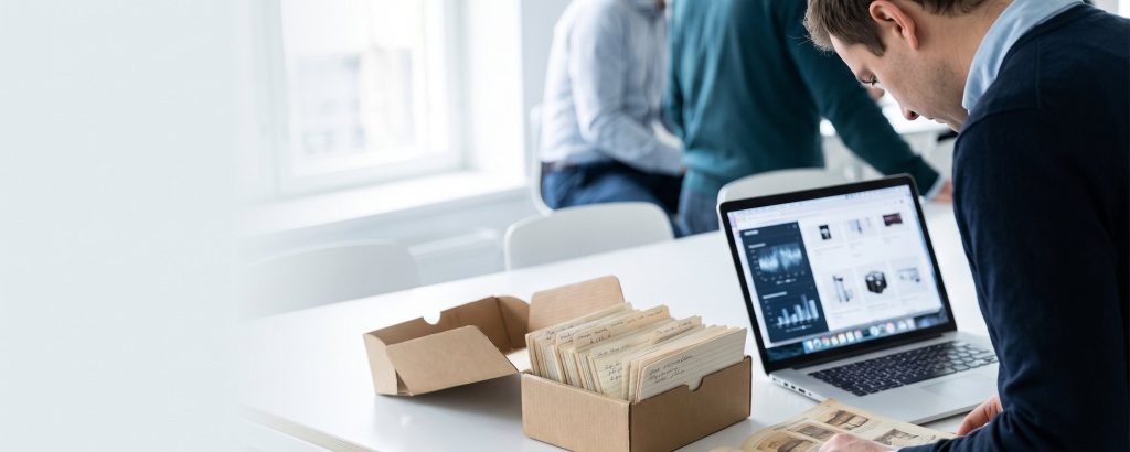 Produktmanager vergleicht alten Katalog und Karteikarten mit Produktdaten am Laptop