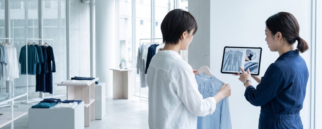 Kundin mit Bluse, Verkäuferin zeigt Produktdetails auf dem Tablet