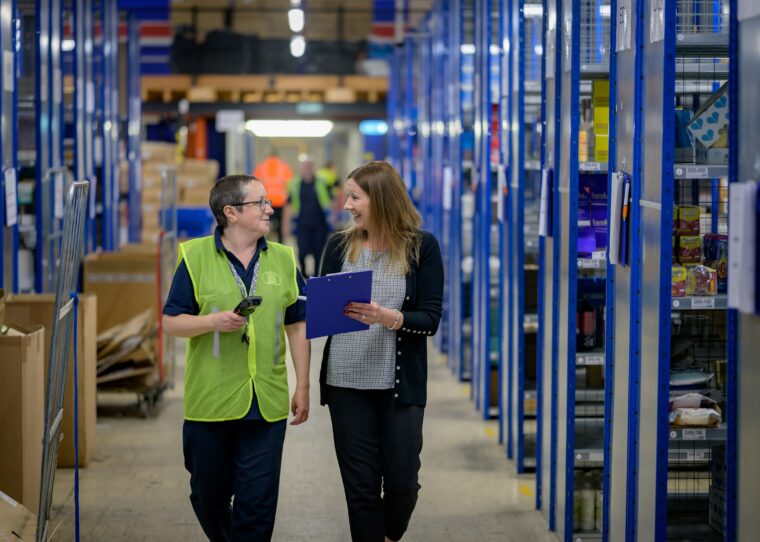 Two employees with a barcode scanner and clipboard at organised warehouse shelving — structured product data at apollon